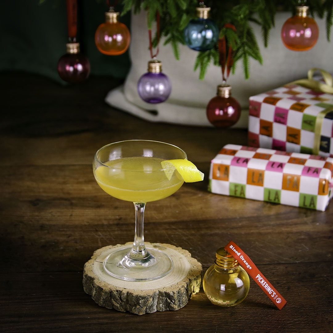 Cocktail in a glass with a lemon twist on a wooden coaster, surrounded by Christmas decorations.