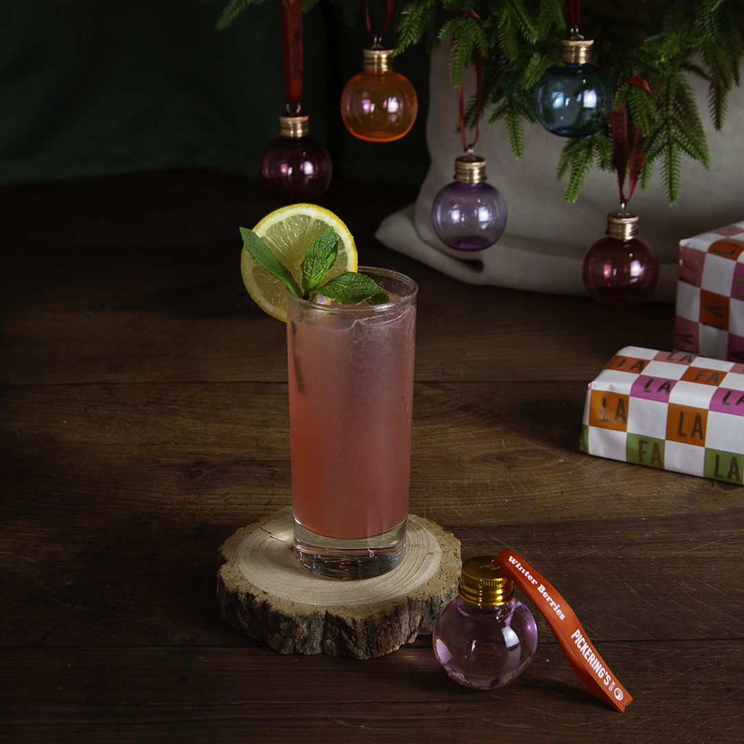 Pink cocktail with lime and mint garnish on a wooden coaster, Christmas decorations in the background.